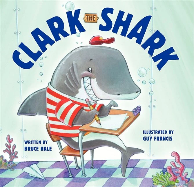 In the center, a large, friendly gray shark named Clark wears a red-and-white striped t-shirt and a small red baseball cap balanced on his head [Image Context]. He has a wide, enthusiastic grin showing rows of sharp white teeth. Clark is squeezed into a small, brown wooden school desk with metal legs, sitting on a matching wooden chair. He holds a yellow pencil in his fin and is eagerly writing on a piece of paper on his desk.
